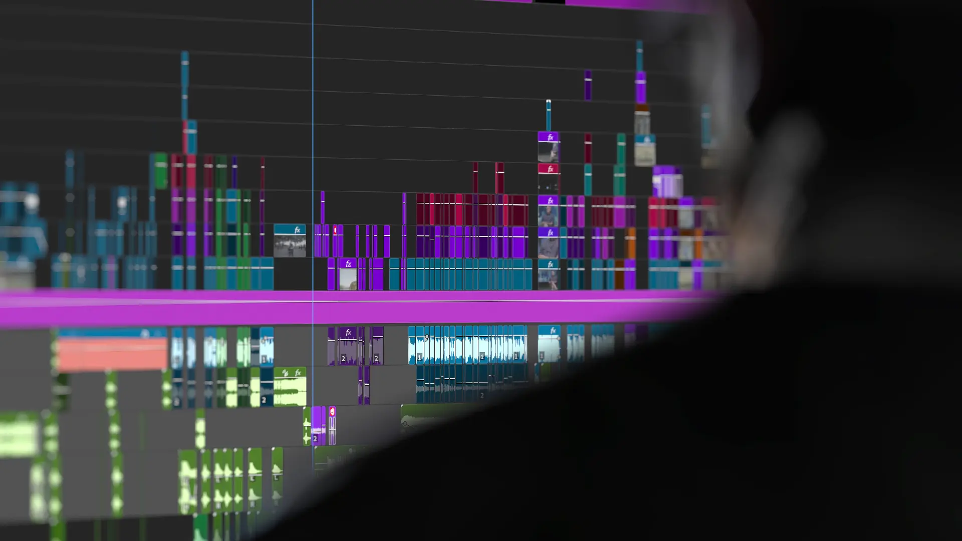 A blurred person faces a computer screen displaying a colorful, complex video editing timeline with multiple tracks and layers in shades of purple, green, and blue.