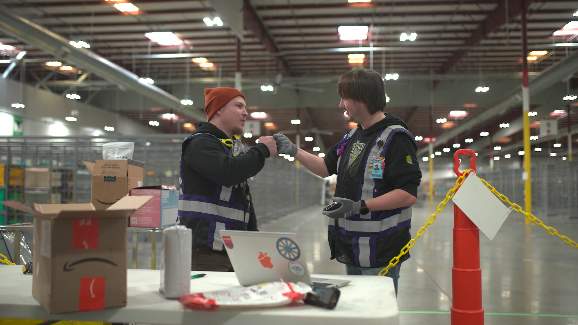 Two warehouse workers in safety vests and gloves bump fists and smile at each other near packages in a spacious, well-lit California facility—the final leg of delivery for Amazon Rural Super Rural documentary brand video capturing the process and spirit on the floor.