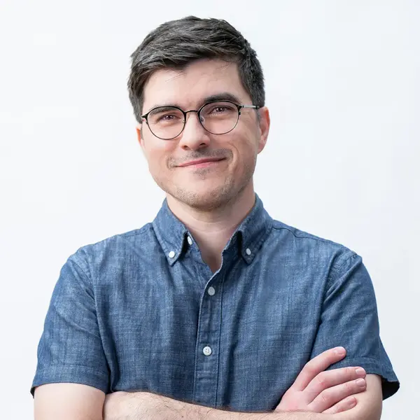 Ben Smith, Art Director, with short brown hair and glasses, wearing a blue button-up shirt, stands with arms crossed and smiles at the camera against a plain white background.