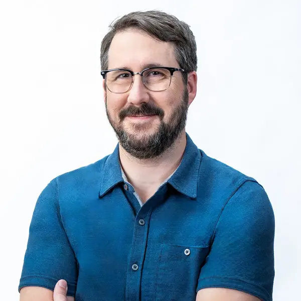Chris Valente, Executive Producer, with short brown hair, a beard, and glasses, is smiling with arms crossed. He wears a blue shirt and stands confidently against a plain white background.