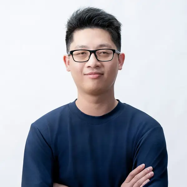 Cleon Cen - Editor, a young man with short black hair and glasses, is wearing a dark blue shirt, smiling slightly, and standing with his arms crossed against a plain white background.
