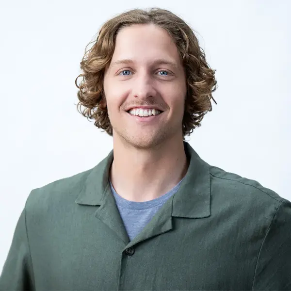 Smiling man with wavy, shoulder-length light brown hair, wearing a green collared shirt over a gray t-shirt, stands in front of a plain light background. Kyle Chater - Staff Designer.