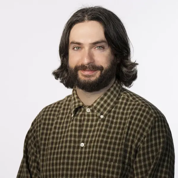 Logan Shillinglaw IV, Senior Editor, with shoulder-length dark hair and a beard, is wearing a green and brown checkered button-up shirt. He is smiling and posed against a plain white background.