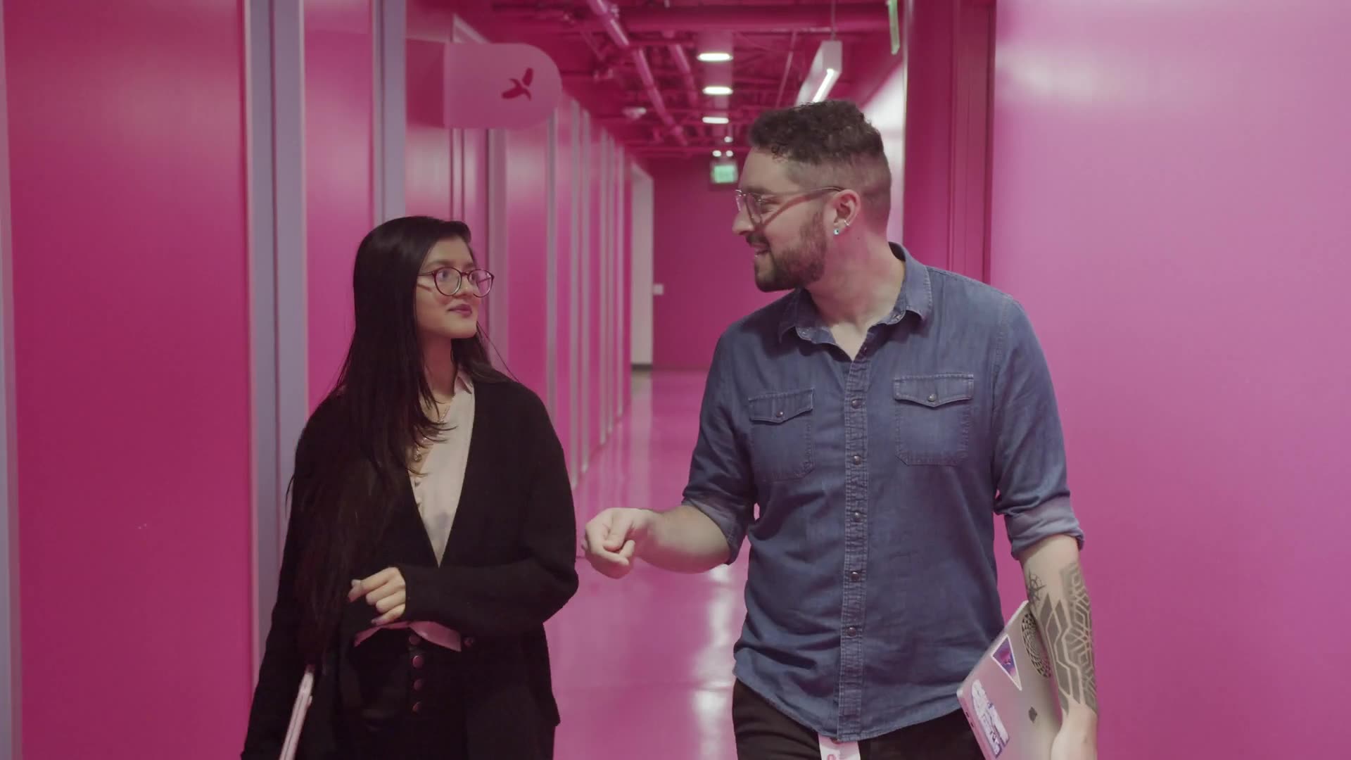 Two people walk side by side in a vibrant pink hallway, engaged in conversation. Filmed for a Lyft Recruitment culture documentary video, one holds a laptop and gestures while the other listens attentively, carrying a bag and folder.