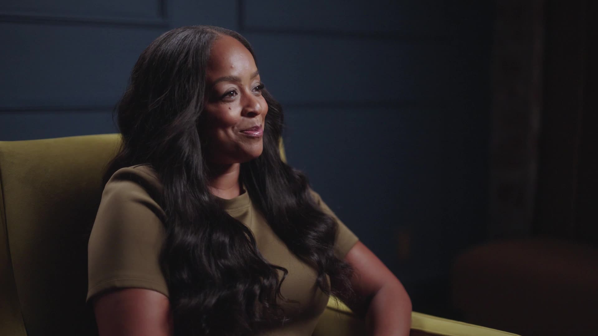 A woman with long, wavy black hair sits in a yellow chair, smiling slightly and looking to the left. She is wearing an olive green top, set against a dark blue wall—perfect for a CreatorIQ SaaS product launch video backdrop.