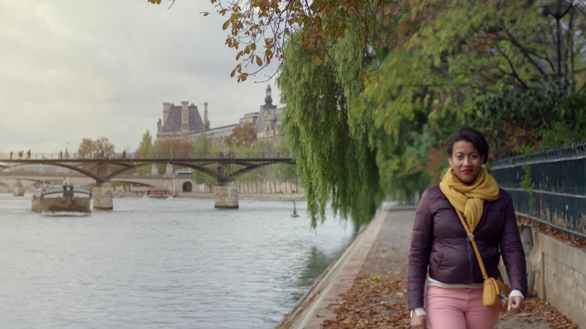 A woman walks along a leaf-strewn path beside the Seine River in Paris, with a bridge and historic buildings in the background on an overcast day, capturing the cosmopolitan feel of a Sage Multilingual brand video.