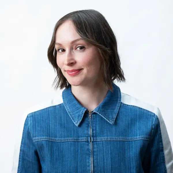 Samantha Sartor - Producer/Director, with short brown hair and a light complexion, smiles softly in a blue denim jacket with a zipper. A white background is behind her.