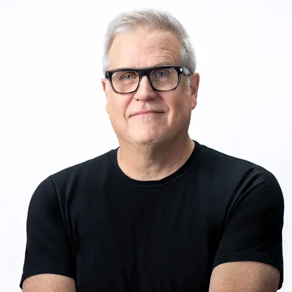 An image of Scott Compton - Director/Founder, a middle-aged man with short gray hair and glasses, wearing a black t-shirt and smiling slightly while facing the camera against a plain white background.