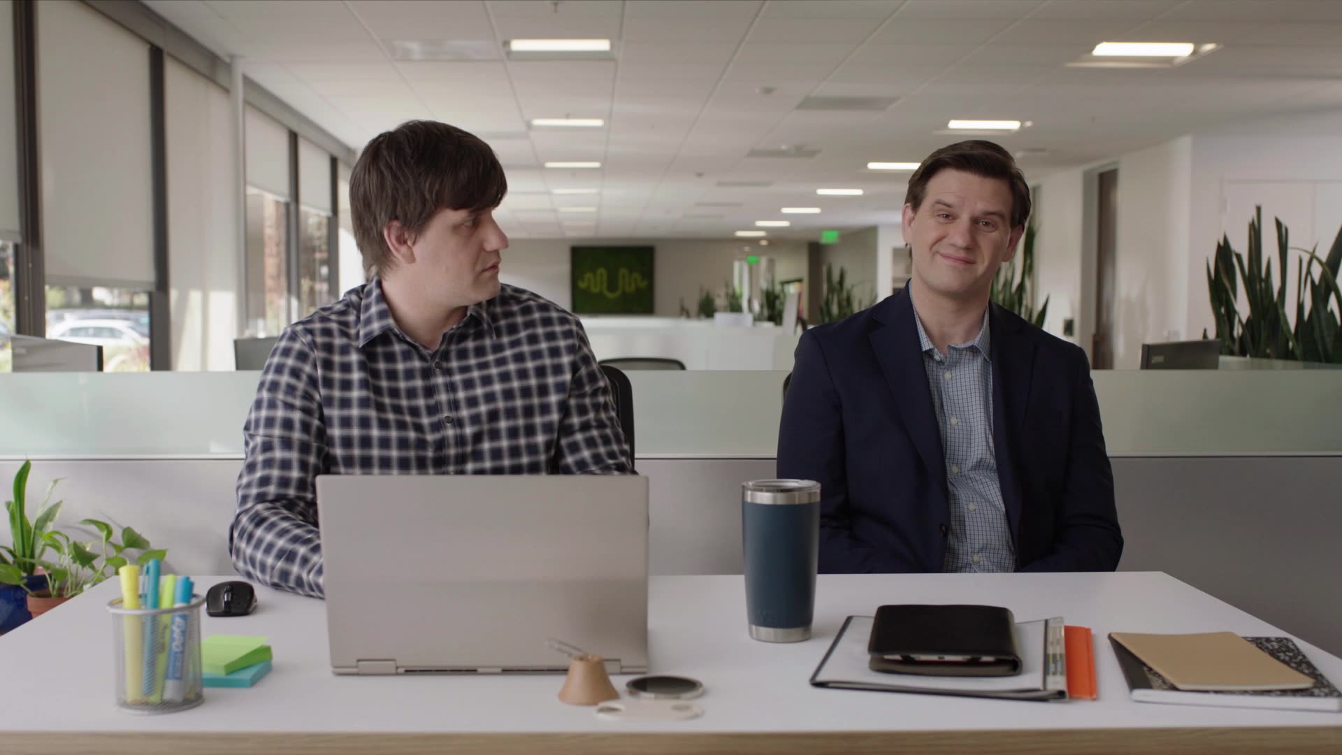 Two men sit at a desk in a modern office working on a brand video. One, in a checked shirt, types on an Amazon laptop while the other smiles. Office supplies and plants are on the desk, with an open, bright workspace behind them.