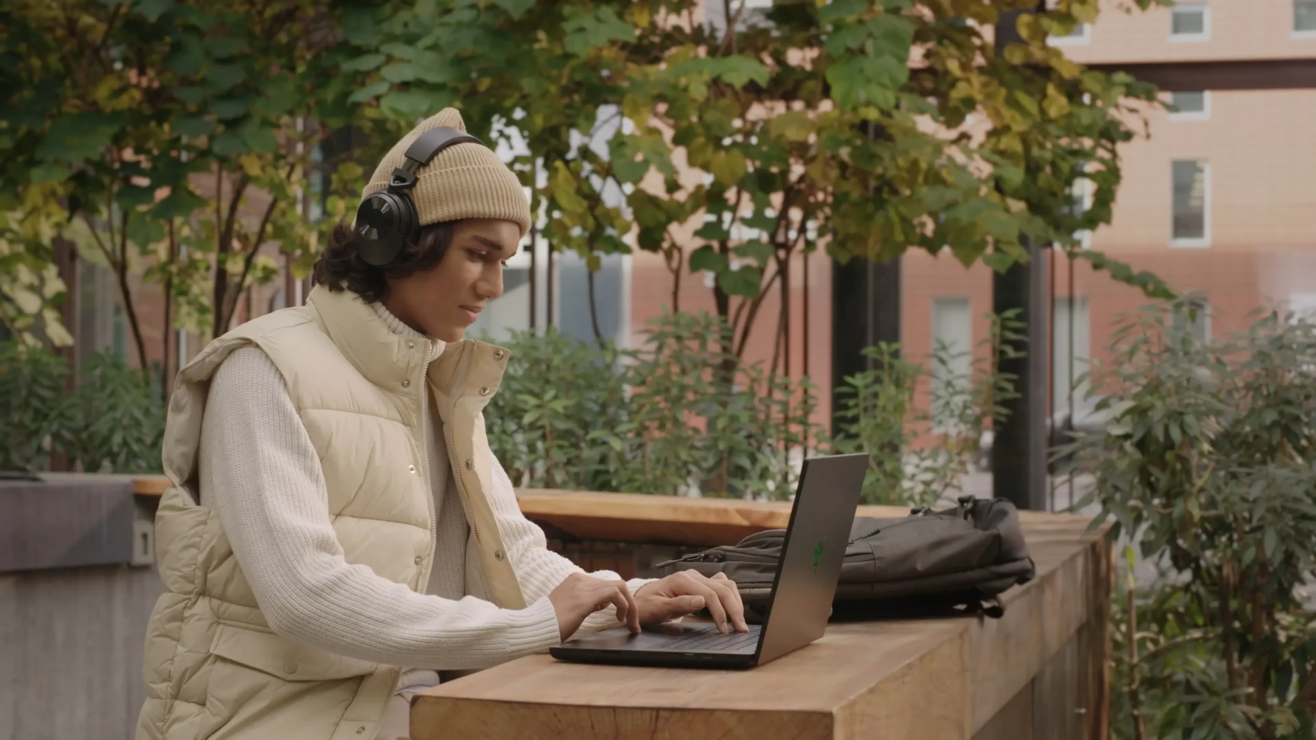 A person wearing a beige beanie, headphones, and a cream-colored vest works on a laptop at an outdoor wooden table, surrounded by greenery and modern buildings—the image is a scene from Splash video of Remedy.