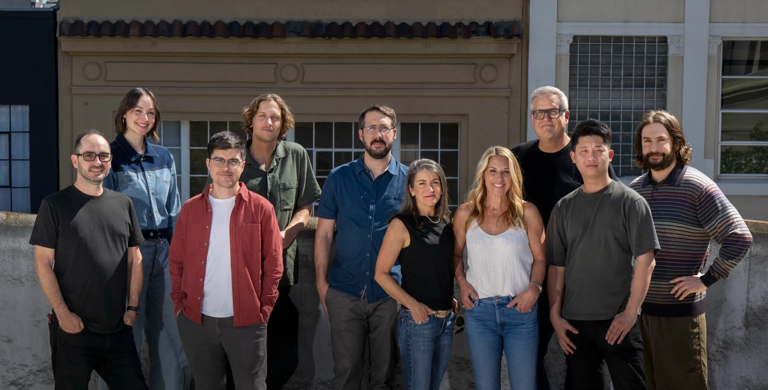 Image of Remedy team: A group of eleven adults stand outdoors in front of a building, smiling and posing for a group photo on a sunny day. The casually dressed Remedy team wears shirts, jeans, and jackets.